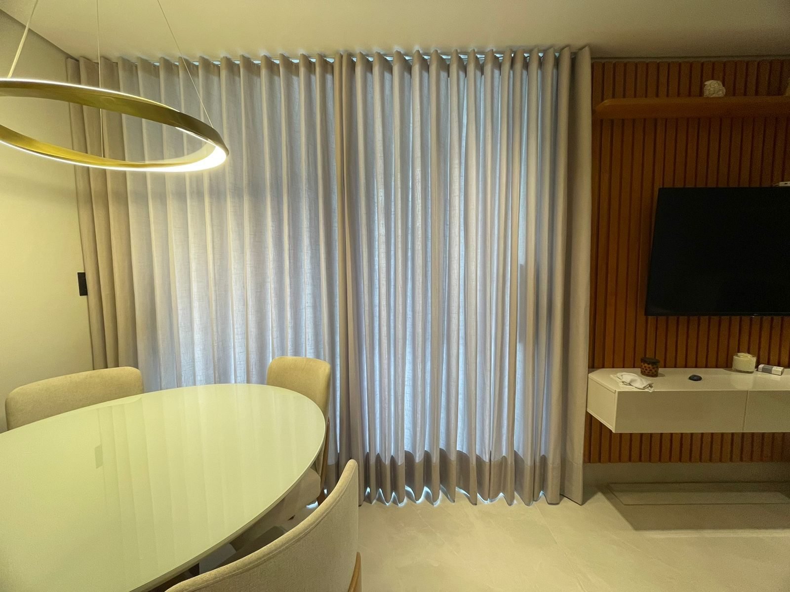 Sala de jantar com pendente circular dourado e cortina wave clara ao lado do painel de madeira
